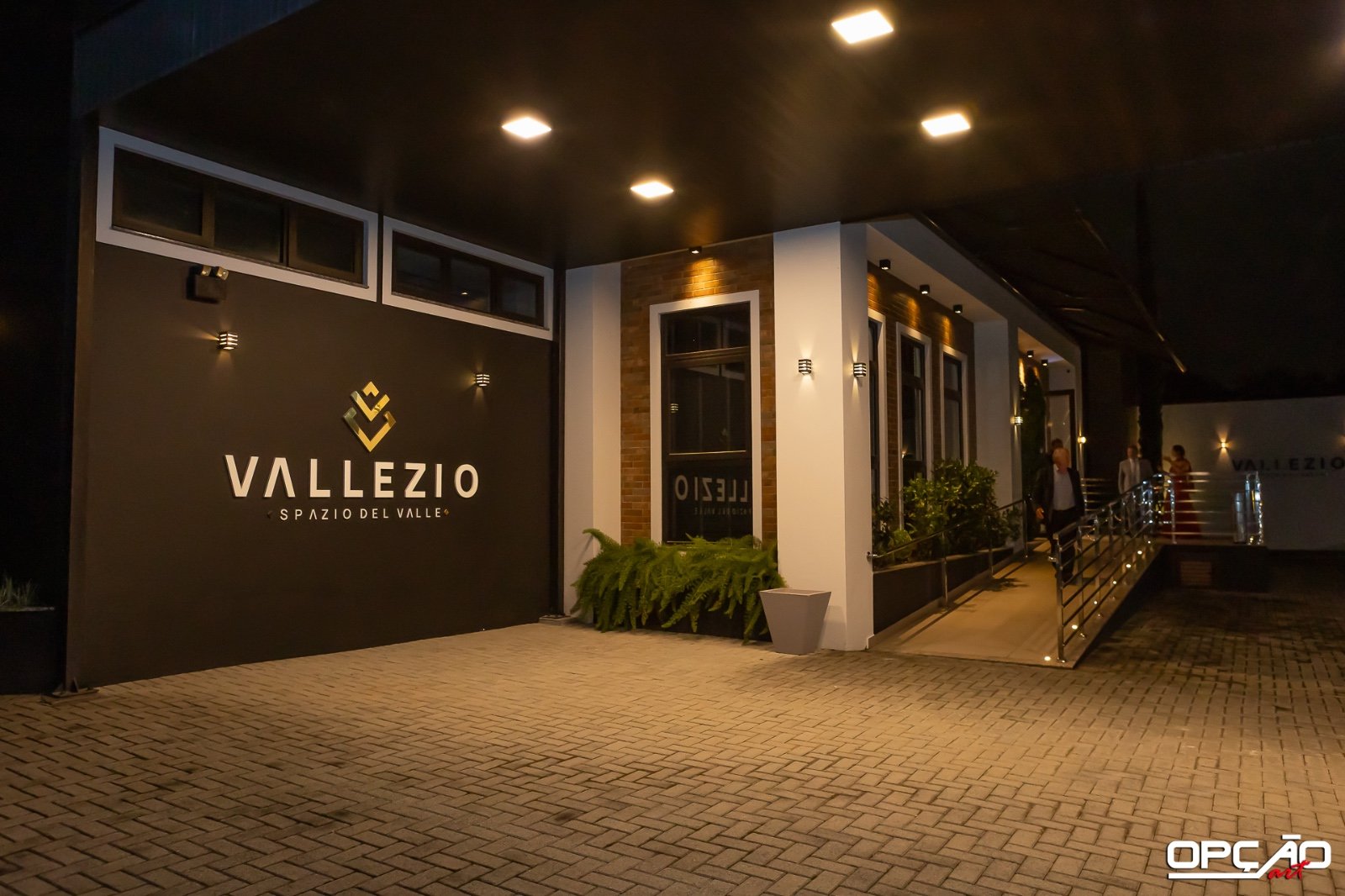 Entrada iluminada à noite, com o letreiro Vallezio Spazio Del Valle e a rampa de acesso